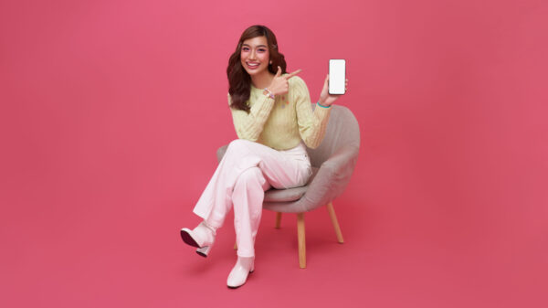 mulher sentada em uma cadeira cinza com a mão direita apontando para o celular que está na mão esquerda. o fundo é rosa
