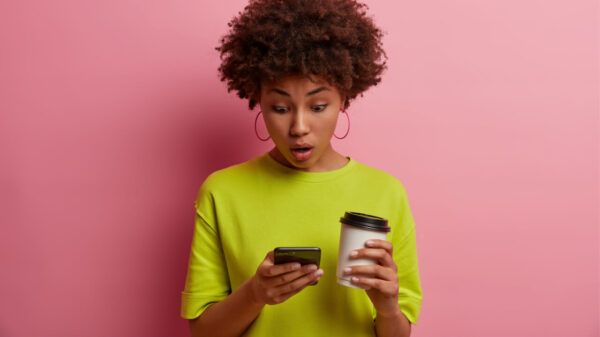 Mulher de cabelos cacheados surpresa, lendo algo no celular, expressando espanto, focada no celular, segurando um copo de café para viagem, isolada em uma parede rosa.
