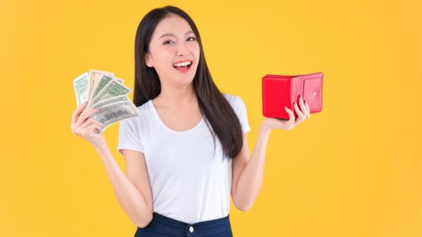 na imagem há uma mulher. na mão esquerda ela segura notas de dinheiro. na mão direita ela segura uma carteira vermelha. o fundo é amarelo