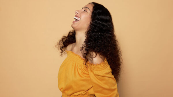 Vista de perfil de uma mulher rindo alto, se divertindo, em pé, contra um fundo bege de um estúdio.