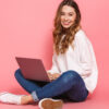 Retrato de uma jovem sorrindo, sentada, com as pernas cruzadas, usando um laptop, isolada sobre um fundo rosa.