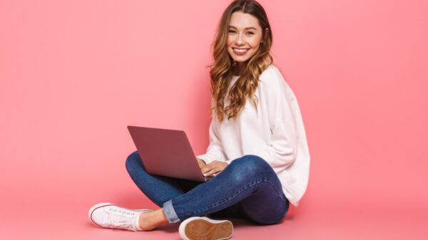 Retrato de uma jovem sorrindo, sentada, com as pernas cruzadas, usando um laptop, isolada sobre um fundo rosa.