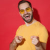 Homem jovem alegre, barbudo, usando óculos casuais e uma camiseta amarela, posando, isolado em um estúdio com um fundo de parede vermelha, apontando os dedos indicadores para a câmera. Conceito de estilo de vida de pessoas.