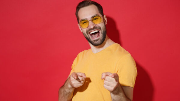 Homem jovem alegre, barbudo, usando óculos casuais e uma camiseta amarela, posando, isolado em um estúdio com um fundo de parede vermelha, apontando os dedos indicadores para a câmera. Conceito de estilo de vida de pessoas.
