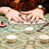 mesa com pano azul, velas e cartas de tarot. sob as cartas está um par de mãos femininas com as unhas pintadas em cor azul