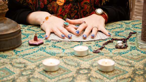 mesa com pano azul, velas e cartas de tarot. sob as cartas está um par de mãos femininas com as unhas pintadas em cor azul