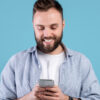 Conceito de vida online. Homem barbudo usando um celular, em um fundo azul de um estúdio. Homem jovem, positivo, com celular moderno, navegando na internet, postando nas mídias sociais.