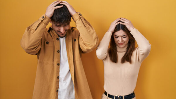 Jovem casal em pé, sobre fundo amarelo, sofrendo, desesperados e estressados, com as mãos na cabeça.