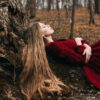 mulher deitada em um tronco de árvore, de cabelos loiros e longos e vestindo um vestido vermelho também longo