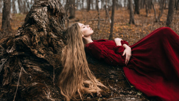 mulher deitada em um tronco de árvore, de cabelos loiros e longos e vestindo um vestido vermelho também longo