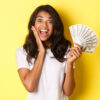 Imagem de uma mulher animada, segurando notas de dinheiro e sorrindo espantada, de pé sobre um fundo amarelo.