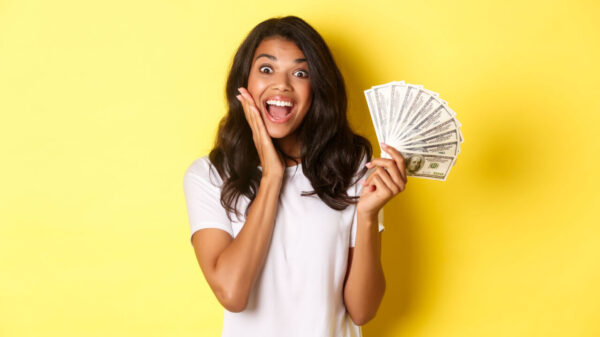 Imagem de uma mulher animada, segurando notas de dinheiro e sorrindo espantada, de pé sobre um fundo amarelo.