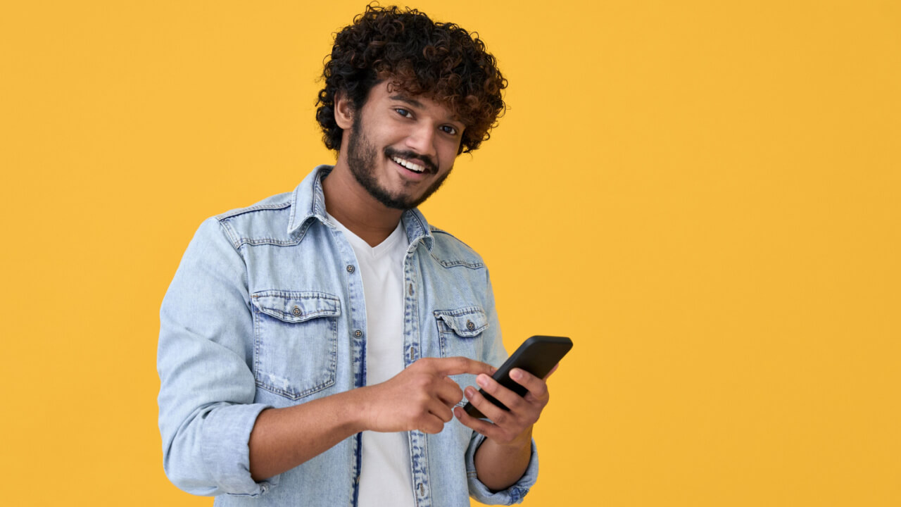 Homem sorrindo, usando um celular, isolado em um fundo amarelo.