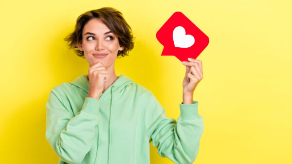 Retrato fotográfico de uma mulher parecendo curiosa, segurando um ícone de coração de mídia social, usando um moletom verde, isolada em um fundo de cor amarela.