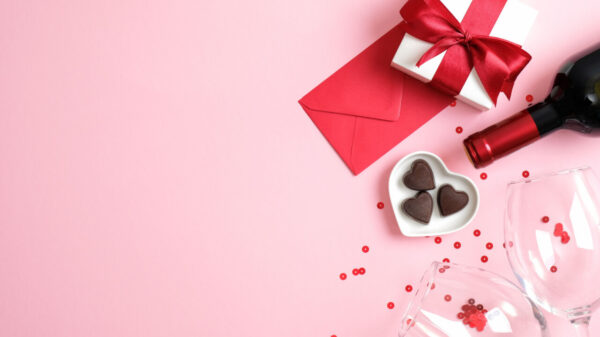 Garrafa de vinho, caixa de presente, copos de vidro, doces em forma de coração, envelope de papel vermelho e confetes, em um fundo rosa. Vista superior.