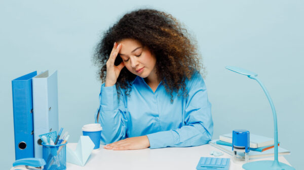 Mulher exausta, triste e cansada, com os olhos fechados, usando uma camisa casual, sentada, com a mão na testa, isolada em um fundo azul pastel. Retrato de estúdio.
