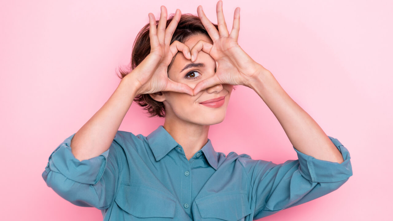 Retrato de uma mulher com os dedos fazendo o formato de um coração na frente de um de seus olhos, isolada em um fundo de cor rosa.