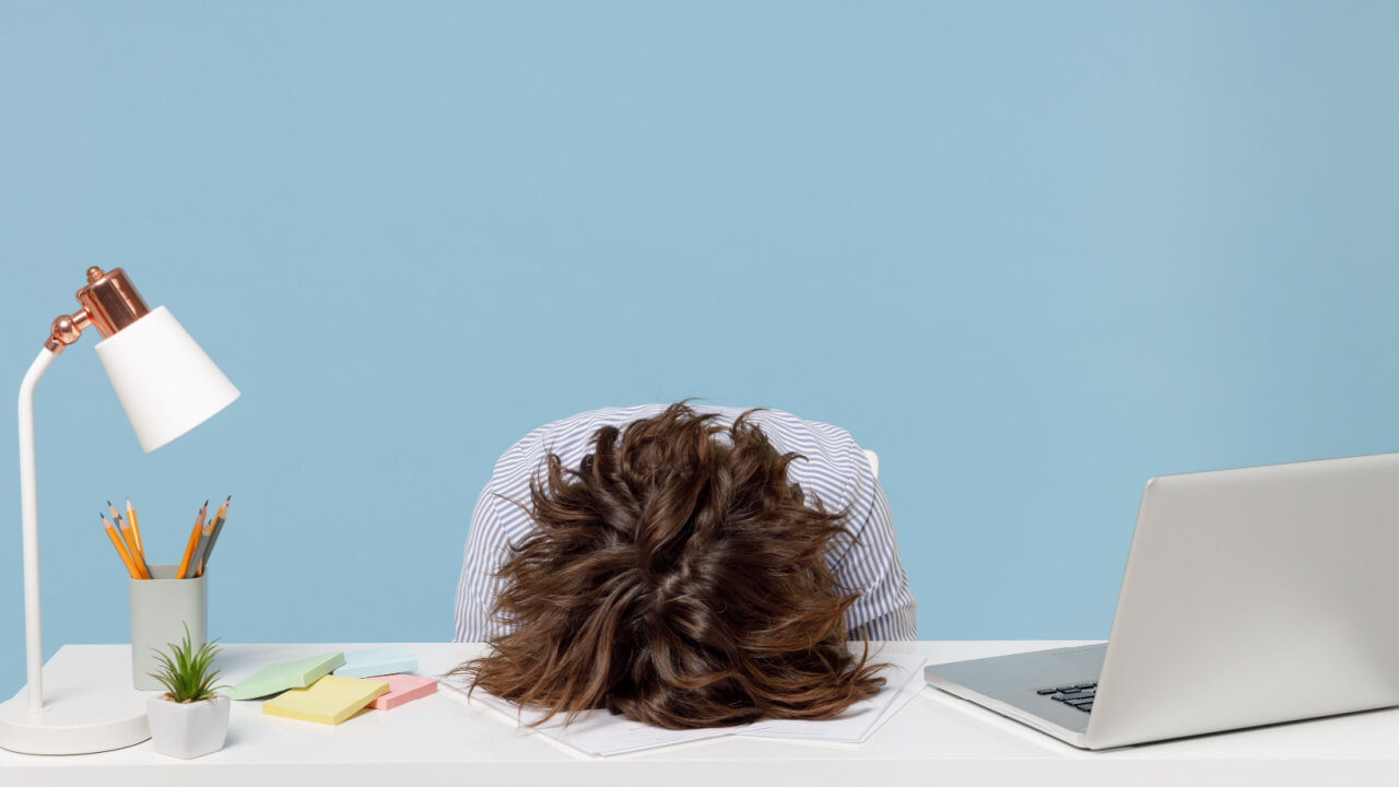 Pessoa cansada, exausta, frustrada, secretária, usando uma camisa casual, sentada, com a cabeça na mesa de escritório branca que contém um laptop, isolada em um estúdio de fundo azul pastel.