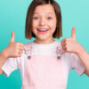 Foto de uma menina alegre e feliz, com os dedos polegares para cima, sorrindo, isolada em fundo de cor azul-esverddeada.