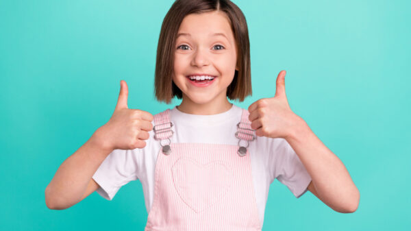Foto de uma menina alegre e feliz, com os dedos polegares para cima, sorrindo, isolada em fundo de cor azul-esverddeada.