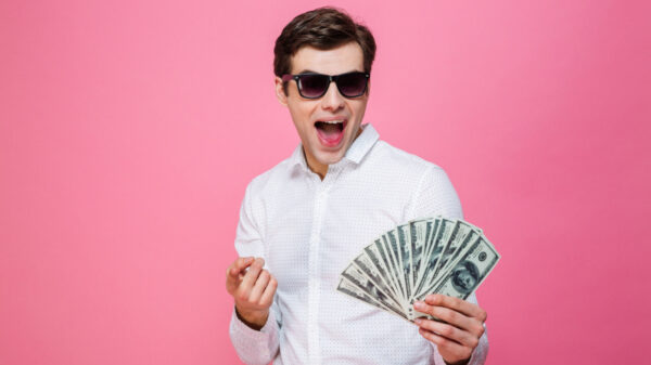 homem segurando um leque de dinheiro, fazendo expressão de felicidade e com óculos escuros em um fundo rosa