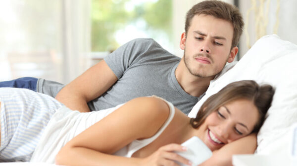 homem olhando para a tela de uma celular por cima do ombro da mulher enquanto eles estão deitados na cama