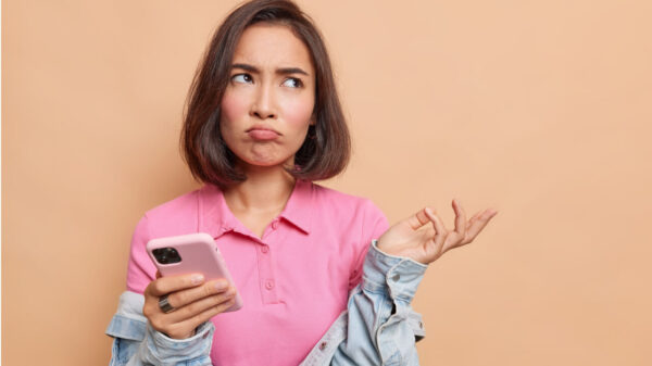 Mulher descontente, com uma expressão de frustração, segurando um celular, usando uma camiseta rosa e uma jaqueta jeans.