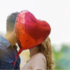 casal ao ar livre se beijando com um balão em formato de coração cobrindo os rostos