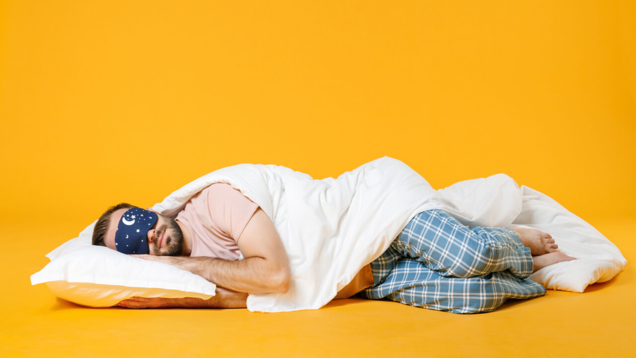 homem dormindo com cobertas e faixa para olhos em fundo amarelo