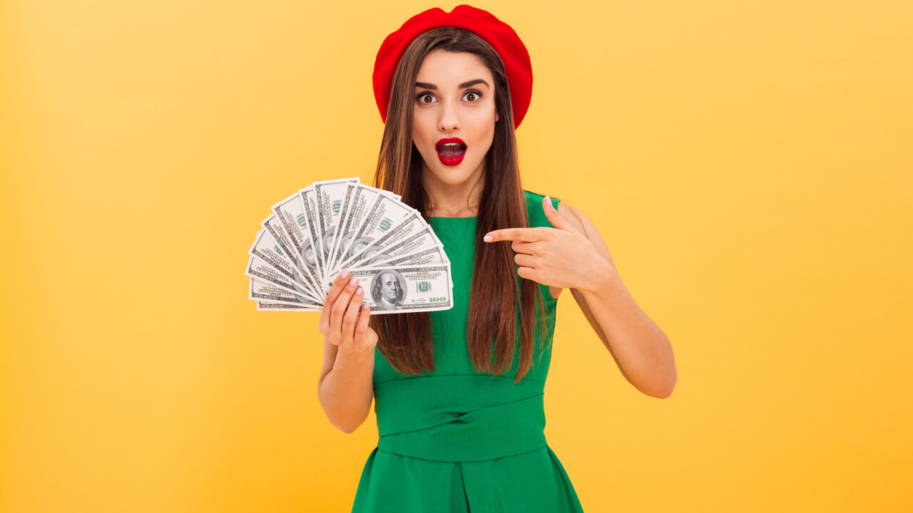 Foto de uma mulher chocada, isolada sobre um fundo amarelo, segurando e apontando para notas de dinheiro, olhando para a câmera.