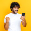 Homem feliz e animado segurando o celular, em um fundo laranja isolado, com uma expressão facial alegre, sorrindo, gesticulando com o punho.