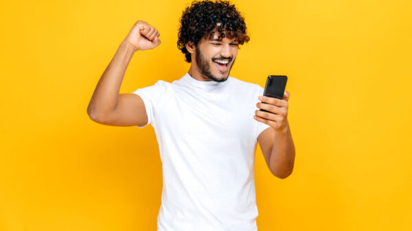 Homem surpreso e animado, segurando smartphone, recebendo notícias inesperadas, em um fundo laranja isolado, com uma expressão facial alegre, gesticulando com o punho.