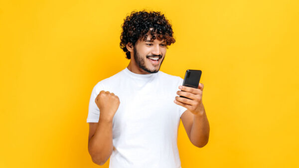 Homem feliz e animado segurando o celular, em um fundo laranja isolado, com uma expressão facial alegre, sorrindo, gesticulando com o punho.