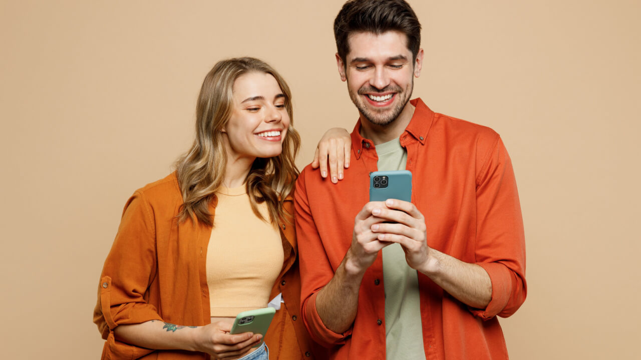Casal de amigos, composto por um homem e uma mulher, feliz, sorrindo, usando roupas casuais, segurando cada um um celular na mão, isolado em um fundo de cor bege clara. Retrato de estúdio.