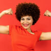 Mulher posando sobre um fundo vermelho, mostrando os dois bíceps, com expressão de incentivo ou motivação.
