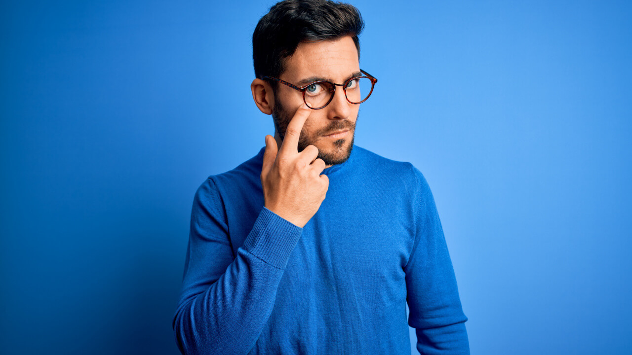 Homem com barba vestindo um suéter casual e usando um óculos, sobre um fundo azul, apontando para o próprio olho.