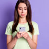 Retrato fotográfico de uma mulher triste e chateada, segurando e olhando para o celular, isolada em um fundo de cor roxa.