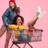 Mulher jovem animada usando uma roupa casual, olhando para a câmera e empurrando um carrinho de compras que está com uma amiga dentro gritando, enquanto as duas se divertem juntas contra um fundo rosa.
