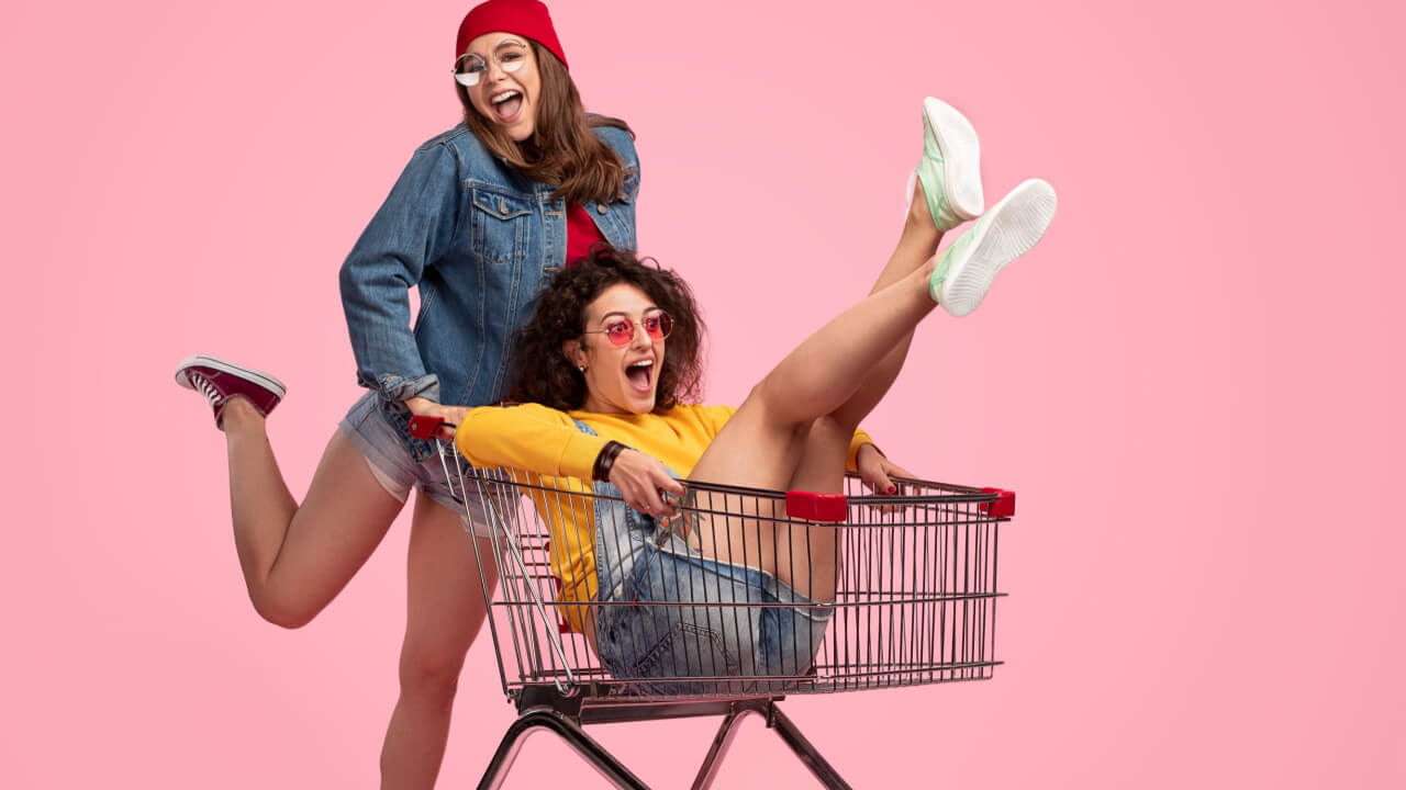 Mulher jovem animada usando uma roupa casual, olhando para a câmera e empurrando um carrinho de compras que está com uma amiga dentro gritando, enquanto as duas se divertem juntas contra um fundo rosa.