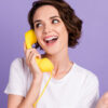 Foto de uma mulher sociável e sorridente falando com alguém em um telefone retrô amarelo, isolada em um fundo de cor roxa.