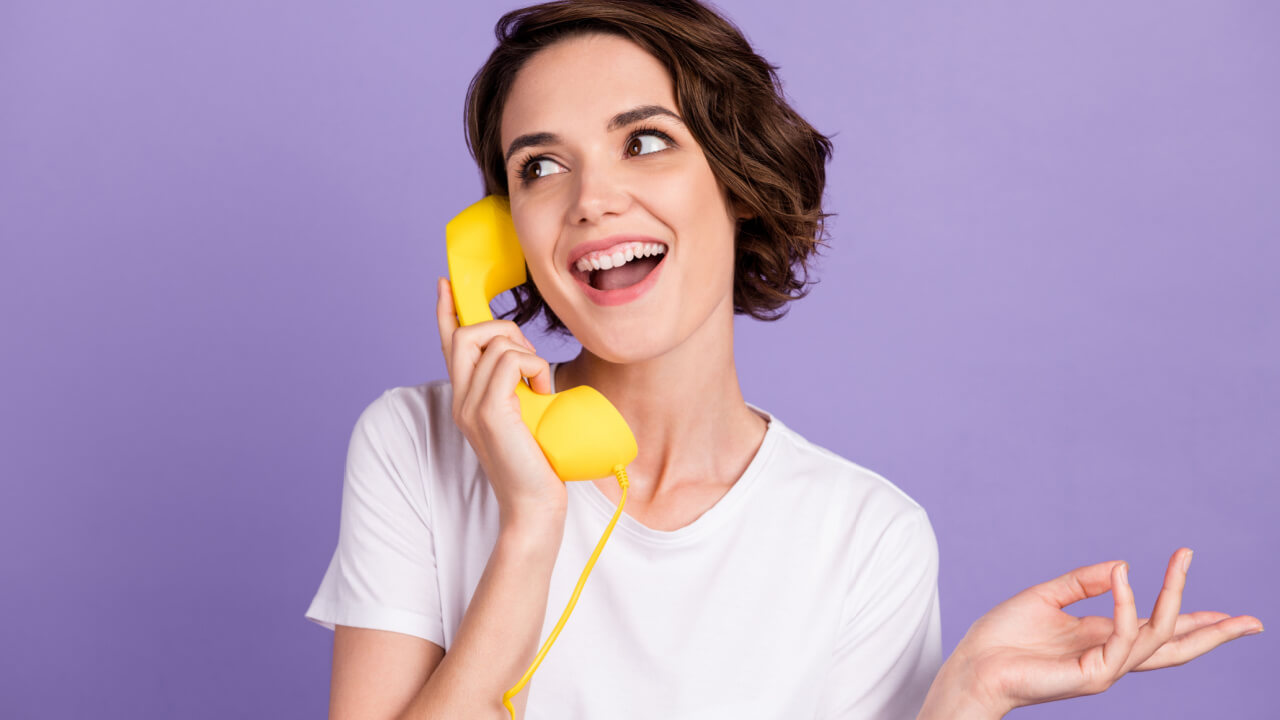 Foto de uma mulher sociável e sorridente falando com alguém em um telefone retrô amarelo, isolada em um fundo de cor roxa.
