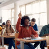 mulher negra sentada em uma sala de aula com a mão levantada