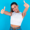 Mulher vestindo uma camiseta branca, sobre um fundo azul, aprovando algo, fazendo gesto positivo com as mãos, com os polegares para cima, sorrindo e feliz.