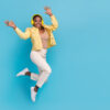 mulher negra pulando feliz vestindo jaqueta e fone de ouvido amarelos em fundo azul