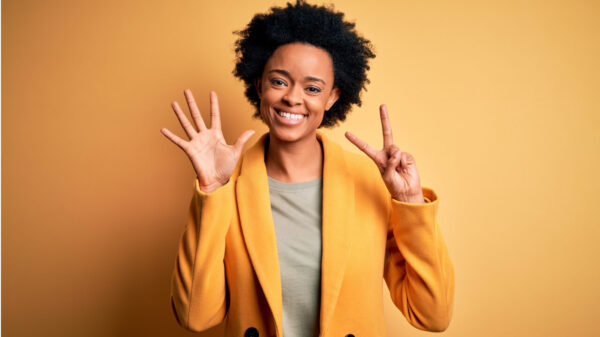 Mulher com cabelos cacheados, vestindo um casaco amarelo, mostrando os dedos fazendo o número sete enquanto sorri confiante e feliz.