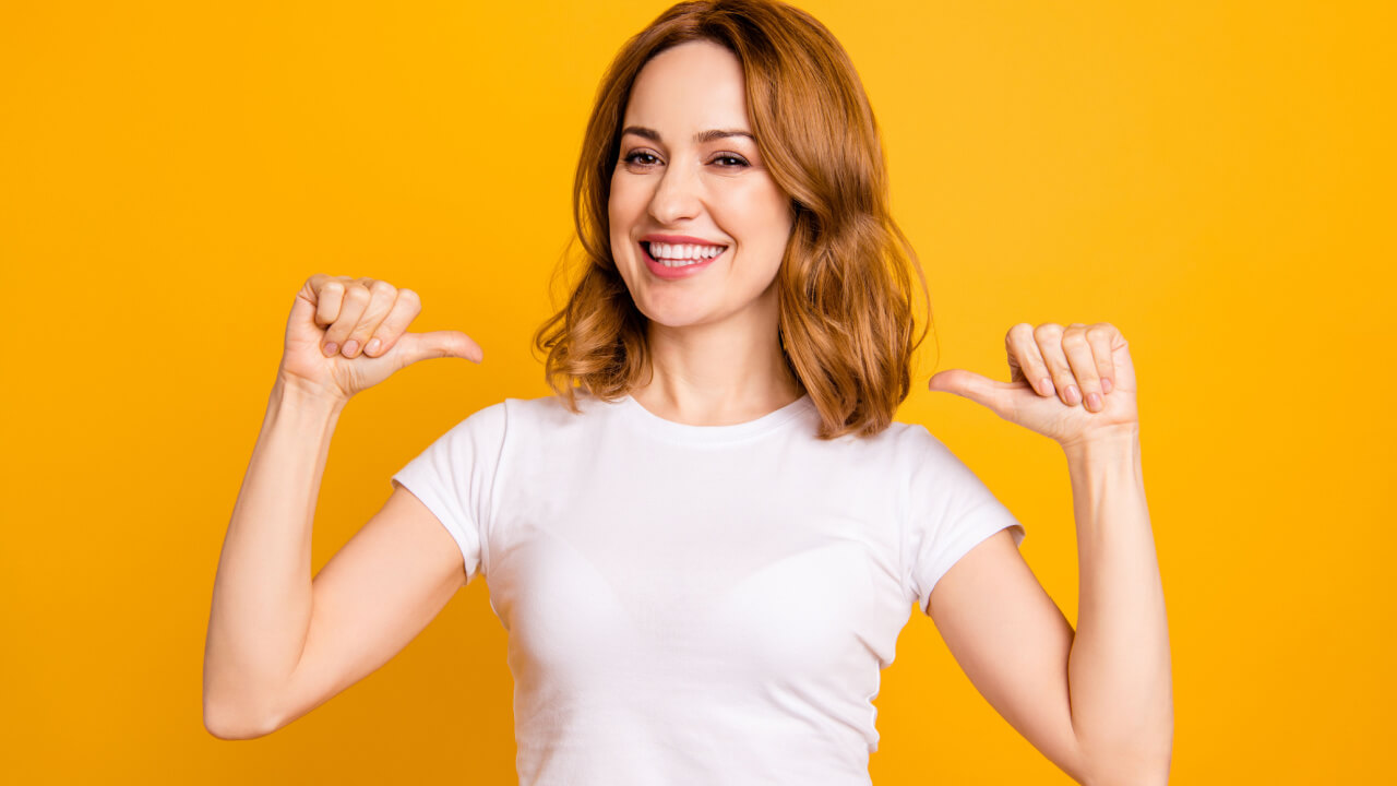 Foto de uma mulher com seus polegares apontados para si mesma, autoconfiante, usando uma camiseta branca casual, isolada em um fundo amarelo.