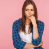Retrato de uma mulher pensativa e chateada, usando uma camisa xadrez, ponderando sobre questões sérias, olhando para o lado com expressão hesitante e incerta. Foto de estúdio interno isolada em um fundo rosa.