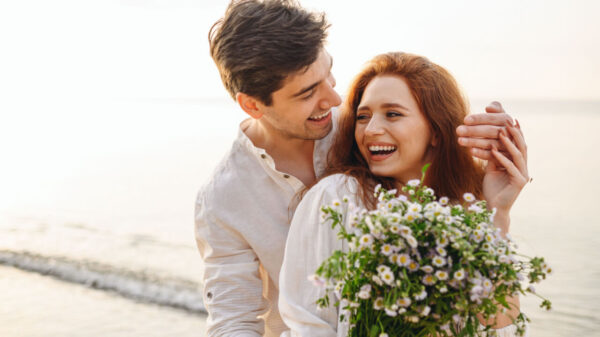 casal heterosexual na praia. os dois usam roupas brancas e estão abraçados. ela segura um buque de flores