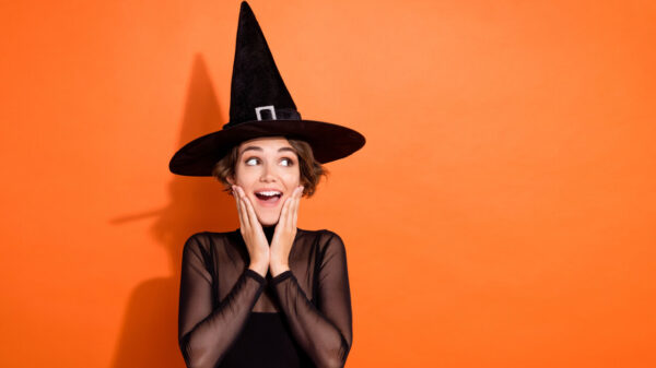 Foto de uma mulher impressionada, usando um chapéu de bruxa preto, com as mãos nas maçãs do rosto, olhando para o lado, em um fundo de cor laranja isolado.