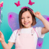 Foto de uma crianças usando asas de borboleta cor-de-rosa, sorrindo, segurando uma varinha mágica, isolada em um fundo de cor azul esverdeado.
